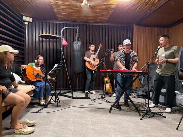 Worship band on keyboard, guitar and vocals in a wood-panelled room while others listen